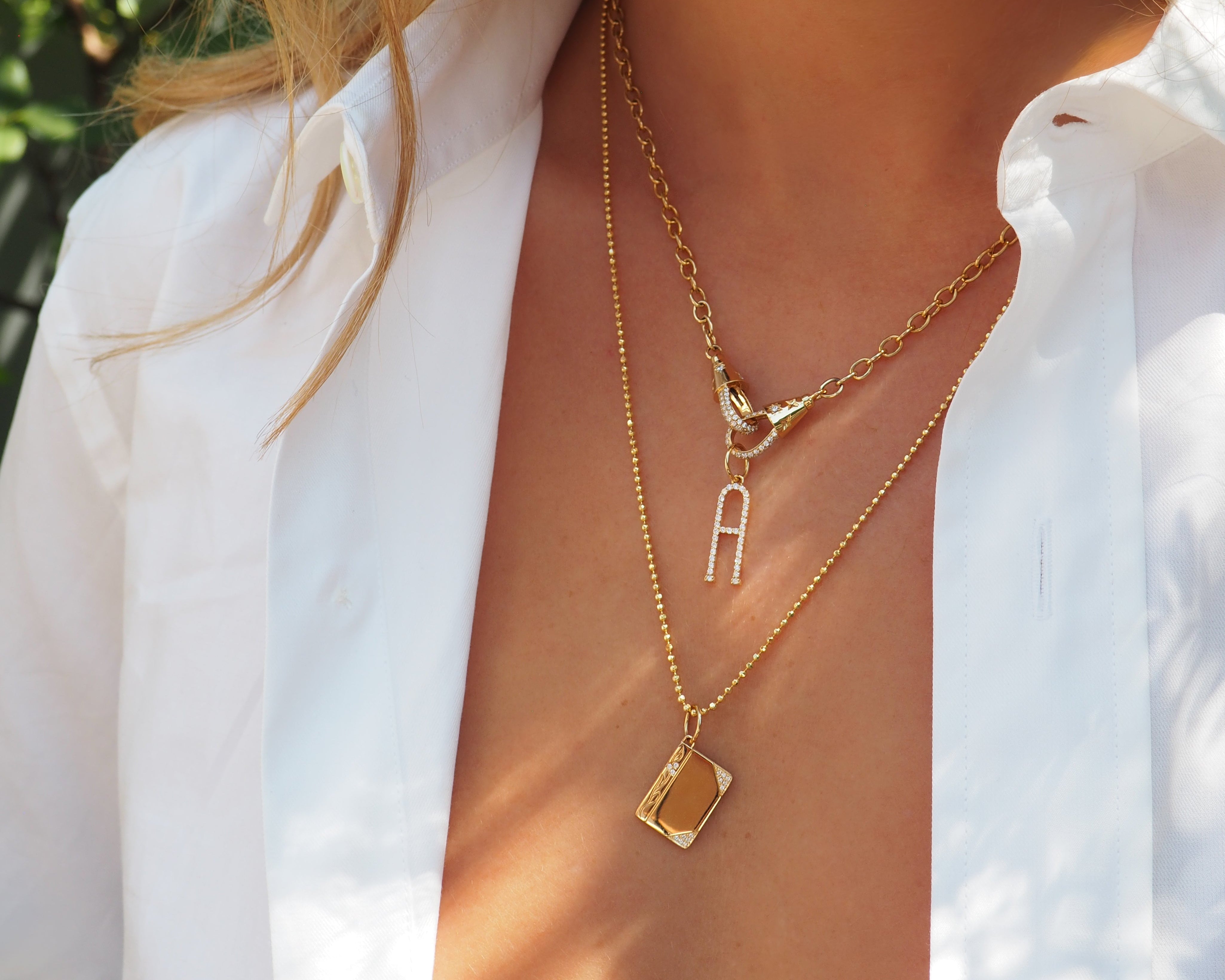 A close-up lifestyle shot of a woman wearing layered gold necklaces, including a delicate chain with a small bird charm and a longer chain with a square pendant, styled with a white collared shirt.