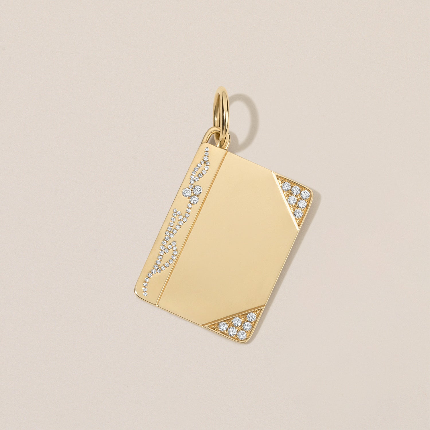 A gold rectangular pendant with a polished finish, featuring pavé-set clear stones along the left edge and bottom corners, shown on a light beige background.